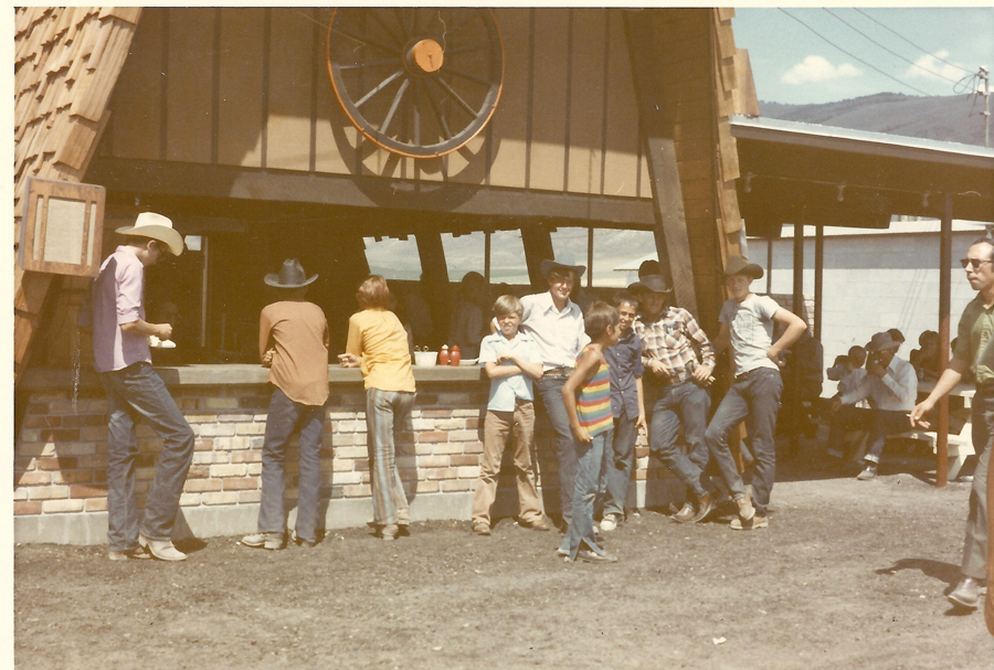 A group of people, mostly wearing cowboy hats, stand around and lean against a brick counter at an outdoor food stand. The counter is part of a building with a large wagon wheel decoration above the service window. Some people are chatting while others look toward the camera. In the background, there are picnic tables with more people seated.