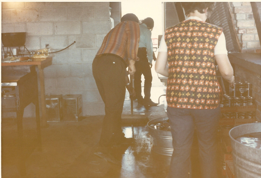 People are in a room with brick and block walls. One person is sweeping the floor, while another stands at a counter. There are various metal buckets and containers, along with tin cans and a bowl on a table.