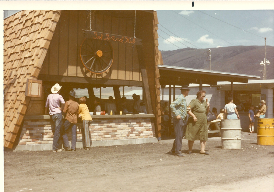 A kiosk with a sign reading "3rd Ward Hut" has people gathered around. Some are standing and talking, while others appear to be ordering or waiting. There is a man in a cowboy hat, and another person wearing a yellow jacket. In the foreground, a man and a woman are walking together. Two barrels are visible nearby. In the background, hills and a partly cloudy sky can be seen.