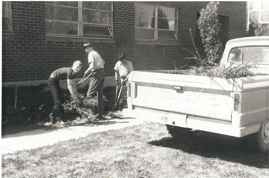 Three people are working outside in front of a brick building with large windows. They are using tools to handle branches and foliage. A pickup truck, with "FORD" written on the back, is parked nearby, carrying more branches. The scene takes place on a grassy area with a sidewalk.