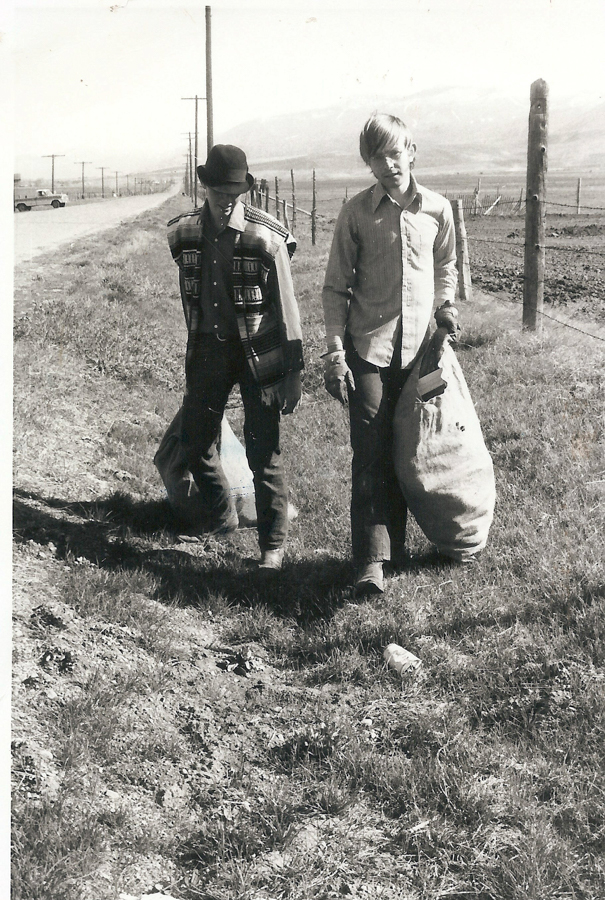 Two boys walking along a rural road, each carrying a large sack. One boy is wearing a hat and a patterned jacket, the other is wearing a button-up shirt. They are on a grassy path next to a field, with mountains in the distance. A truck is visible on the road behind them.