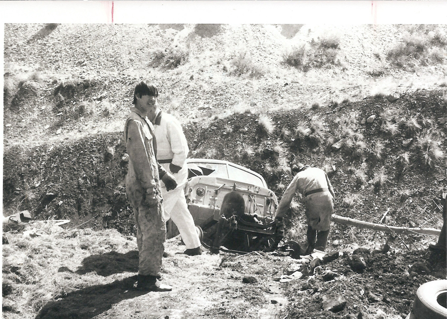 Three individuals in work overalls are near an upturned vehicle in a hilly, grassy area. One person is standing and looking towards the camera, while the other two are examining the vehicle. The vehicle appears to be damaged and is partially on its side. There are various scattered items on the ground around them.