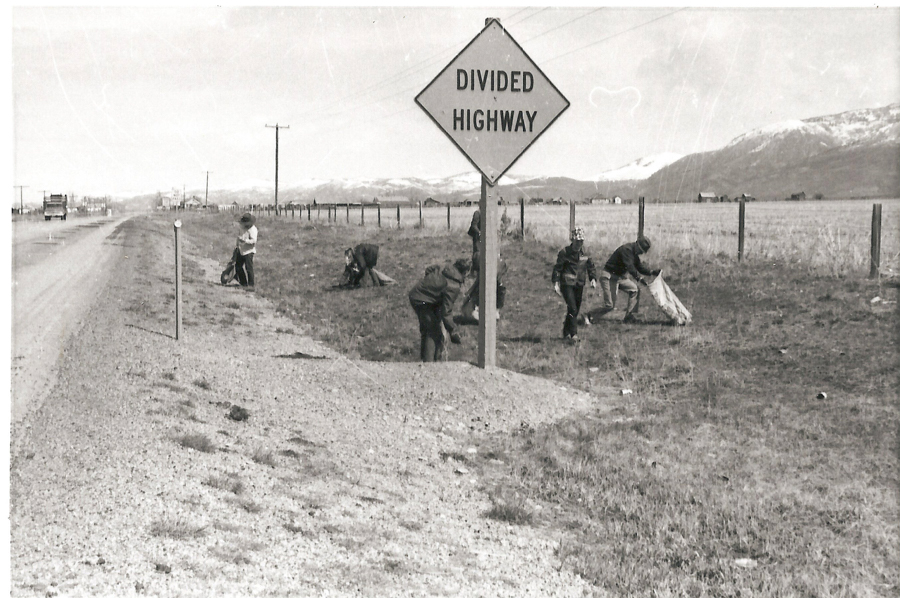 A group of people picking up trash beside a road. They are working near a sign that reads "DIVIDED HIGHWAY." The setting includes grassy fields and mountains in the background. A fence runs alongside the road, and a vehicle is visible in the distance.