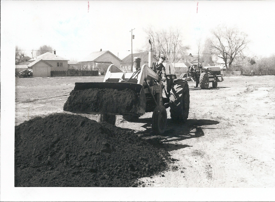 A man operating a tractor with a front loader filled with dirt, positioned next to a large pile of soil. Another tractor is visible in the background. Residential buildings and trees can be seen in the distance.