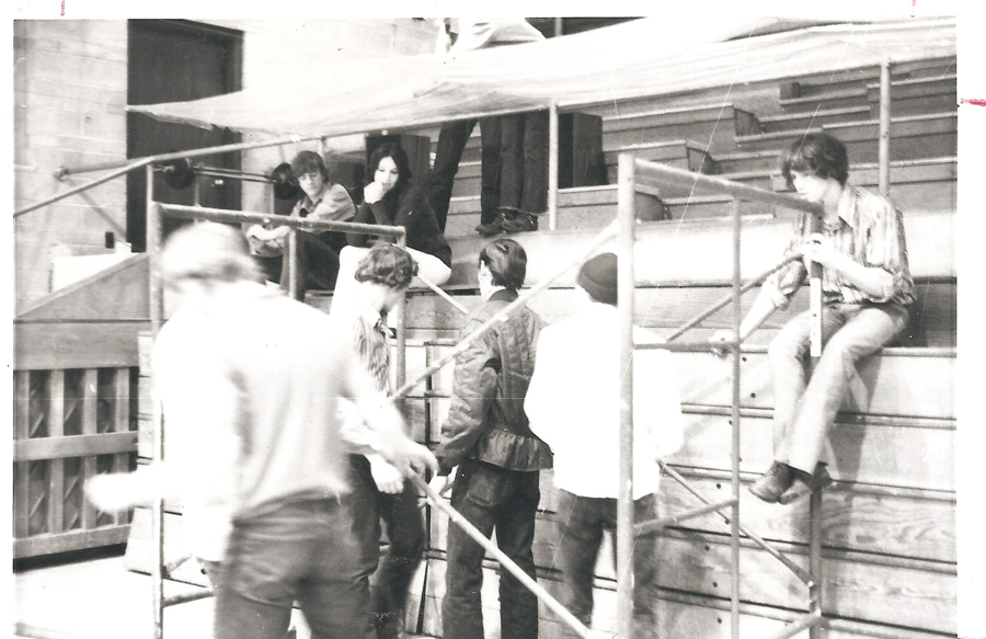A group of people are gathered around scaffolding. Two individuals are sitting on the structure, one on the right holding a piece of it, while several others stand or sit around it. The setting appears to be an outdoor area with steps or seats in the background. Some people are interacting while others are observing.