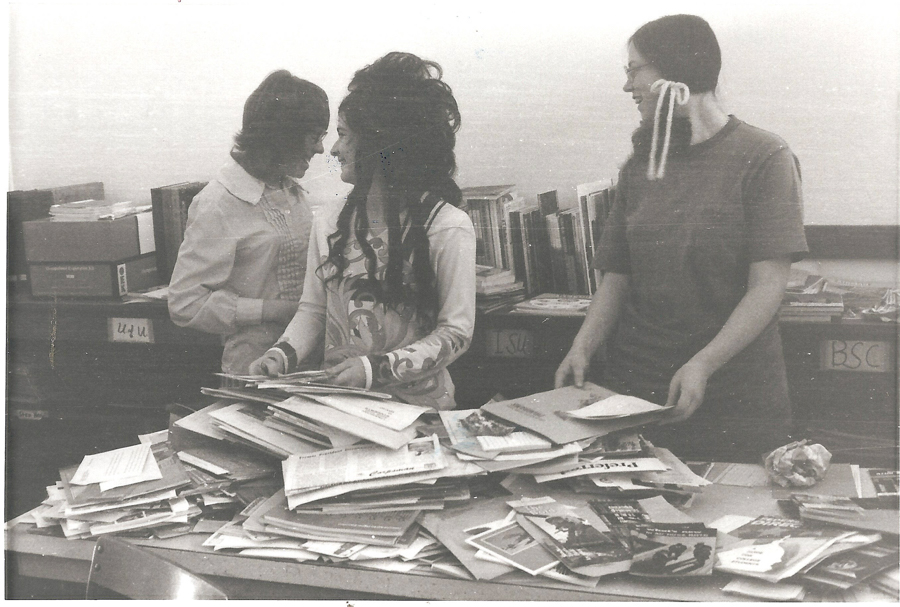 Three people stand behind a table covered in stacks of papers and magazines. The person on the left is wearing a shirt and is facing the other two, who are looking at each other. The person in the middle has long hair and a patterned top, while the person on the right has glasses and a long braid with ribbons. Books and folders are visible on shelves behind them, with labels saying "U.U.," "L.S.U.," and "BSC."