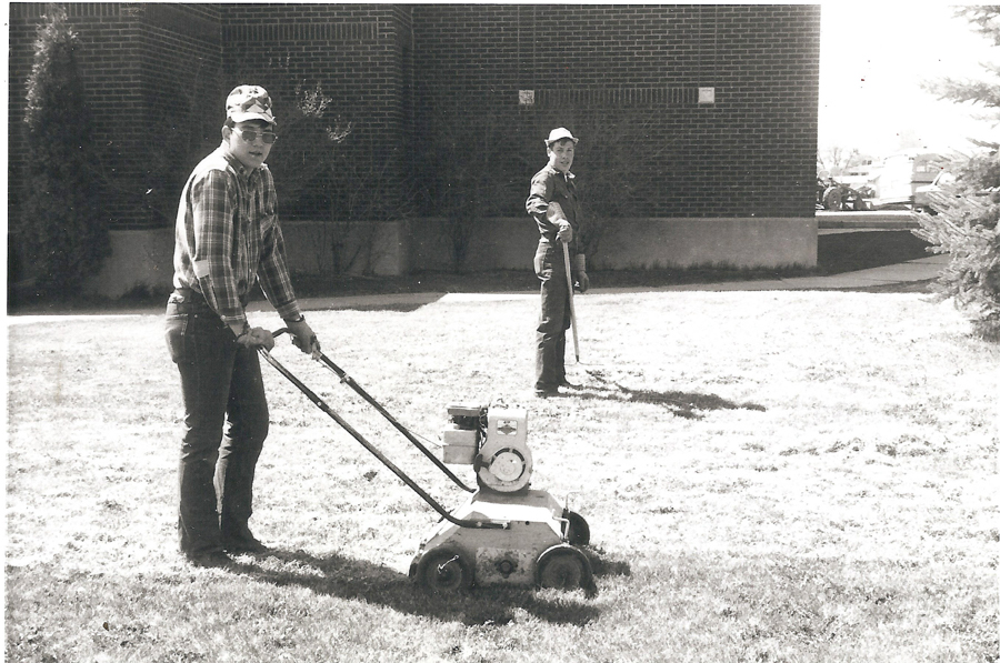 Two men are working on a lawn. One man is pushing a lawn aerator in the foreground, wearing a plaid shirt and cap. The other man, standing in the background, holds a garden tool and is wearing a work outfit and cap. They are near a brick building.