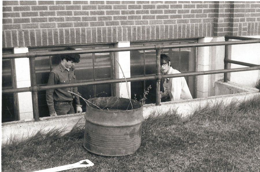 Two people standing in a narrow space between a building and a metal railing. One person is looking down, and the other is looking forward. In front of the railing, there is a large, cylindrical metal container on the ground with some branches or twigs inside. The area is grassy, and a white object lies on the grass. The building has a brick exterior.
