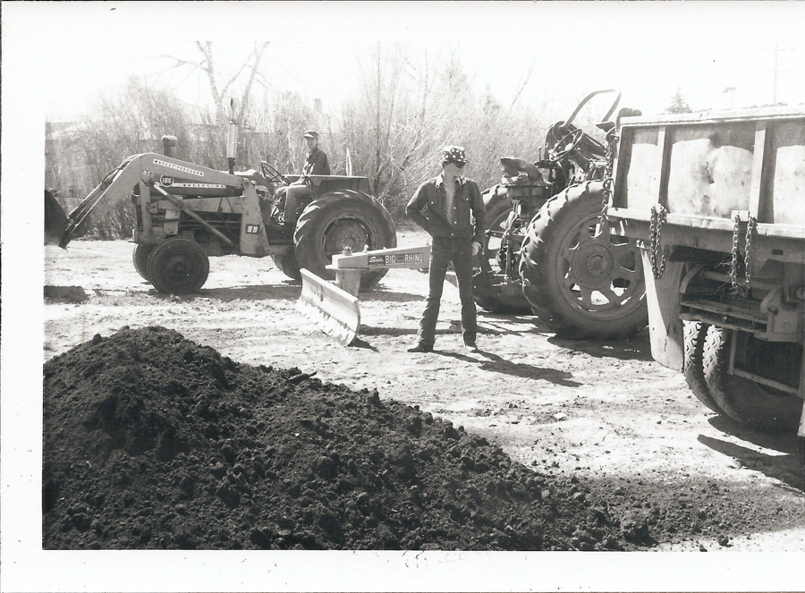 A person stands next to a tractor and a dump truck at a construction site. Another person is seated on a tractor labeled "International 185." The ground is covered in dirt piles, and there are leafless trees in the background. A sign on a plow blade reads "BIG RHINO."
