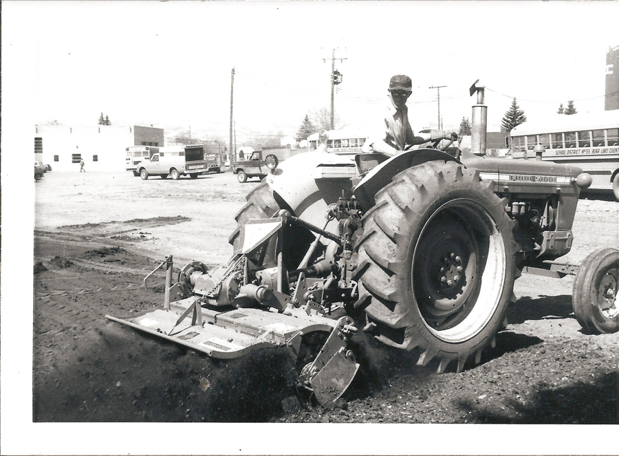 A person driving a large tractor on a dirt surface. The tractor has a plow attachment, and there are buildings, vehicles, and school buses in the background. The text on the bus reads "SCHOOL DISTRICT #33 BEAR LAKE COUNTY." The tractor has the words "FORD 5000" visible on the side.