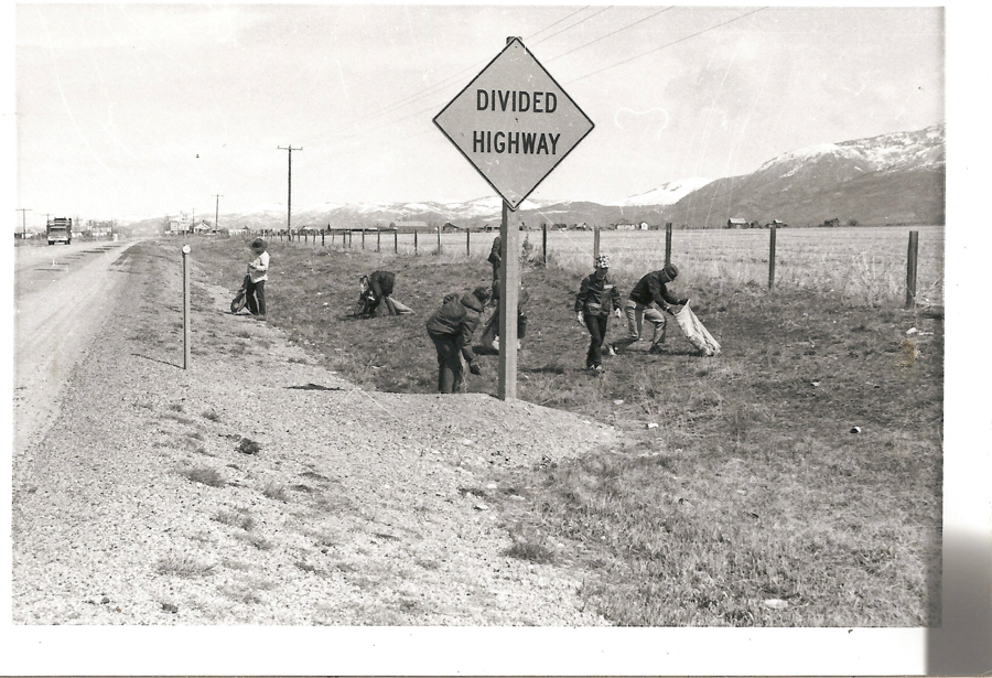 Several people are cleaning up trash along the side of a road. They are collecting litter into bags. A sign nearby reads "DIVIDED HIGHWAY." In the background, there are mountains and a few buildings. A gravel road runs parallel to the fence line.