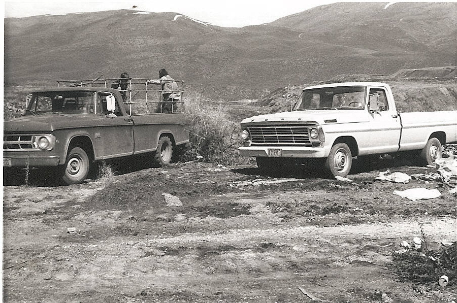 Two pickup trucks are parked on rough terrain with mountains in the background. One truck has a metal frame on the back, with people sitting in the rear. The other truck is parked nearby, with a person sitting inside. The license plate on one truck reads "799921." The ground is uneven and rocky.