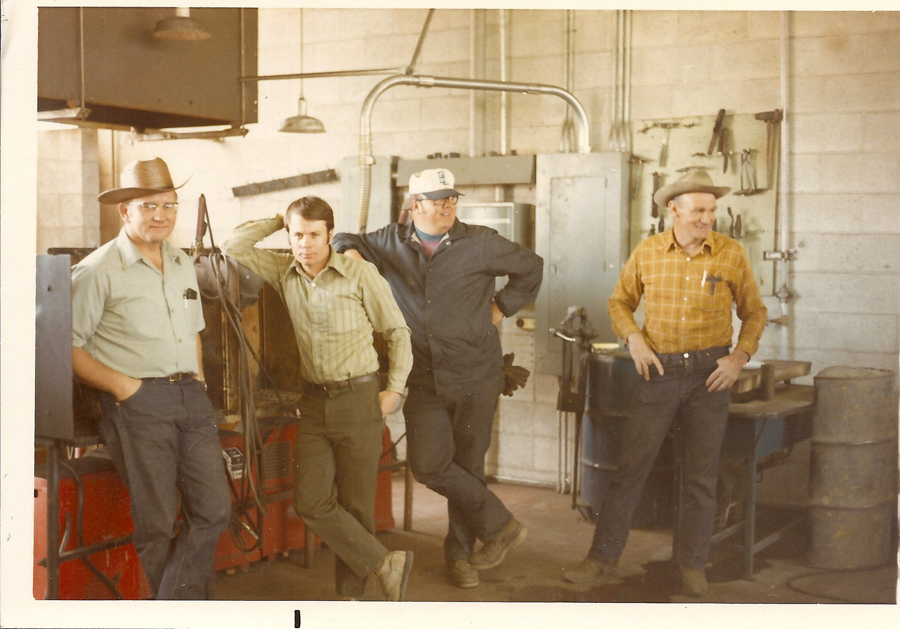 Four men are standing in a workshop. The first man on the left wears a hat and glasses, with his hands in his pockets. The second man leans against a piece of equipment and has his hand on his head. The third man wears a cap, glasses, and work clothes, with one hand on his hip. The fourth man on the right, also wearing a hat, looks to the side and has his hands in his pockets. The background includes tools on the wall, metal barrels, and industrial equipment.