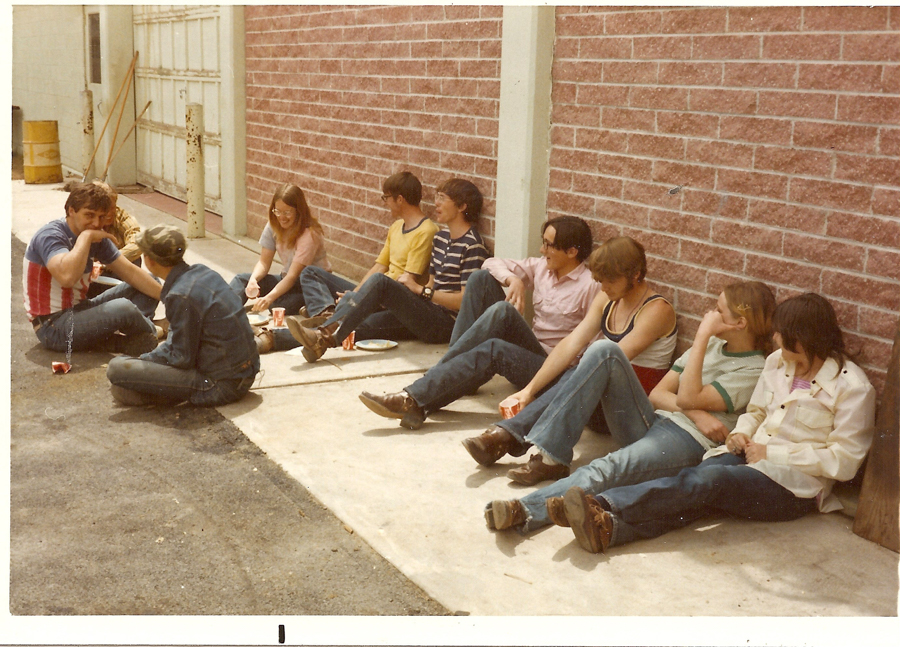 A group of people sitting on the ground outside against a brick wall. They are casually dressed and appear to be having a conversation and sharing food. Some have drinks and paper plates. The setting seems relaxed, and there are items like a barrel and a broom leaning against a nearby wall.