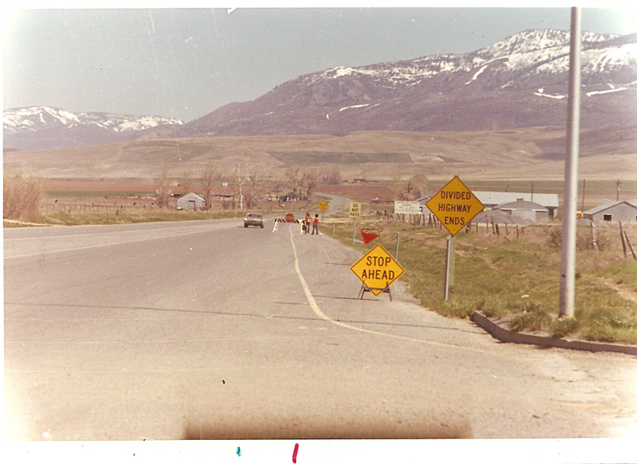 A road with signs reading "STOP AHEAD" and "DIVIDED HIGHWAY ENDS" beside it. In the distance, a car and several people are near some barricades. Snow-capped mountains are visible in the background, with fields and a few buildings nearby.