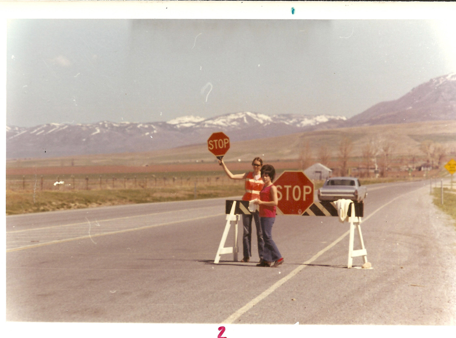 Two people are standing on a road behind a barricade with a stop sign. One is holding a stop sign. There are mountains in the background, and a car is parked on the road.