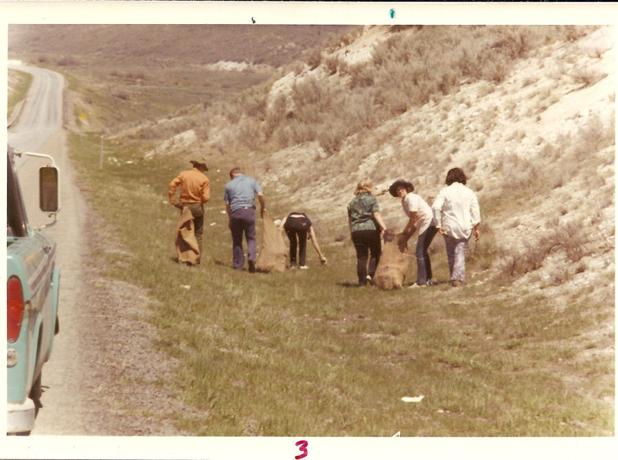A group of people standing near the side of a road, with a hillside on the opposite side. They appear to be picking up something from the ground. One person is bending over, while others are holding or carrying large bags. A truck is parked on the roadside. The number "3" is written at the bottom of the image.