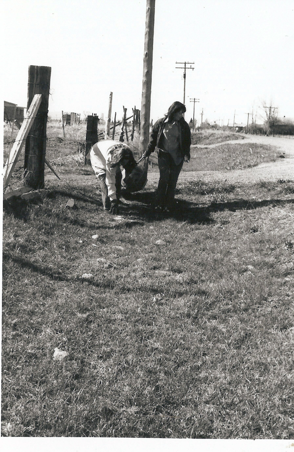 Two people in a field, one bending down with their back turned, holding hands with the other who is standing upright. They are near a wooden fence and utility poles, with a dirt path and open land stretching into the distance.