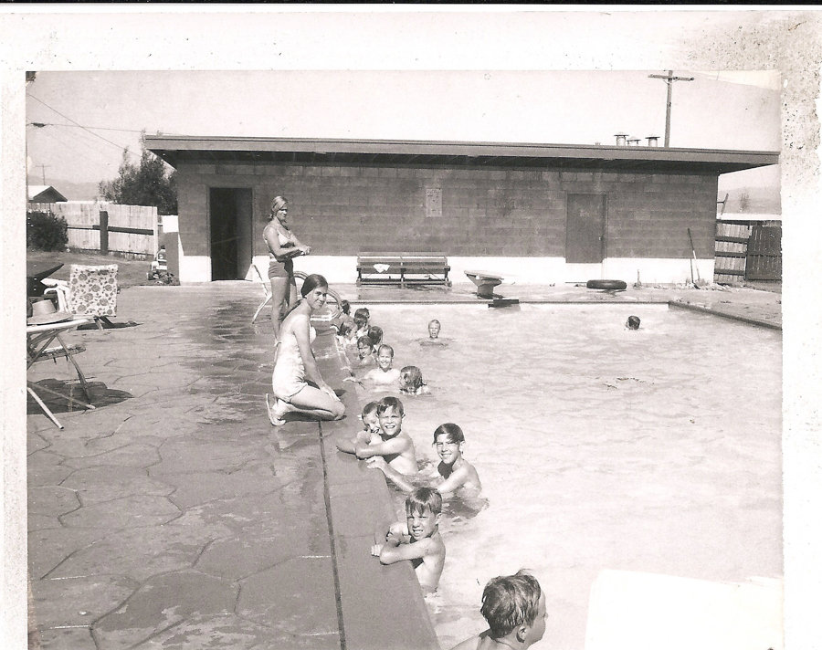 A group of children is swimming in a pool, with a few sitting on the edge. A woman kneels at the poolside, watching the children. Another person stands near a building in the background. There are lounge chairs and a bench around the pool area. The setting appears to be a recreational or community swimming pool.
