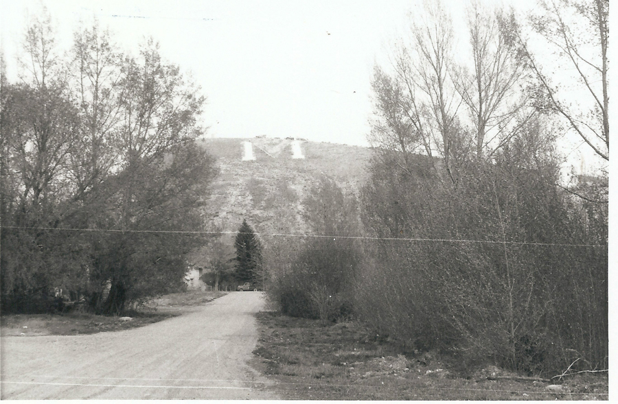 A dirt road leads towards a hill in the background, which has a large letter "M" marked on it. Trees line both sides of the road, and a distant house and vehicle are visible among the trees.