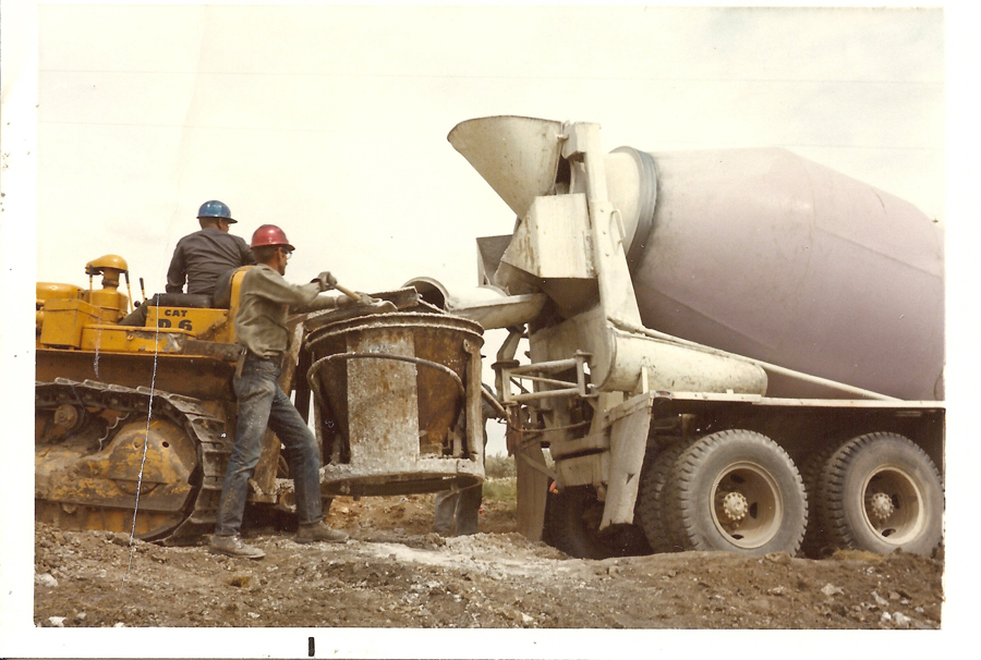Two men wearing helmets and work clothes are operating construction equipment. One man stands near a large metal container suspended from a machine connected to a concrete mixer truck. The other man is seated on a yellow bulldozer with "CAT D6" written on it. The ground is covered in dirt and construction material.