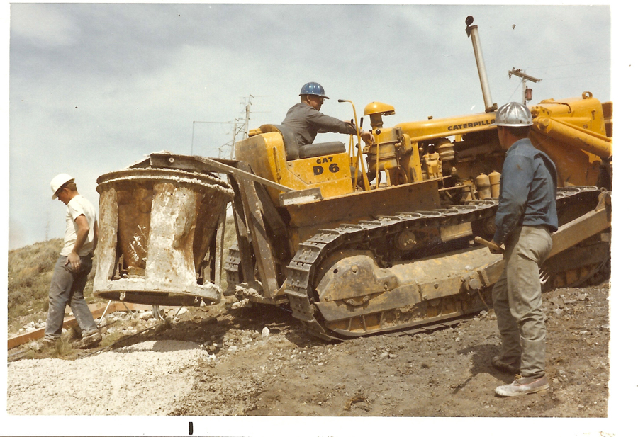 A construction scene with three men wearing hard hats. One man operates a yellow Caterpillar D6 bulldozer, marked with "CAT D 6," while the other two men stand nearby on a dirt and gravel surface. A large metal bucket is attached to the bulldozer. Electric poles and lines are visible in the background.