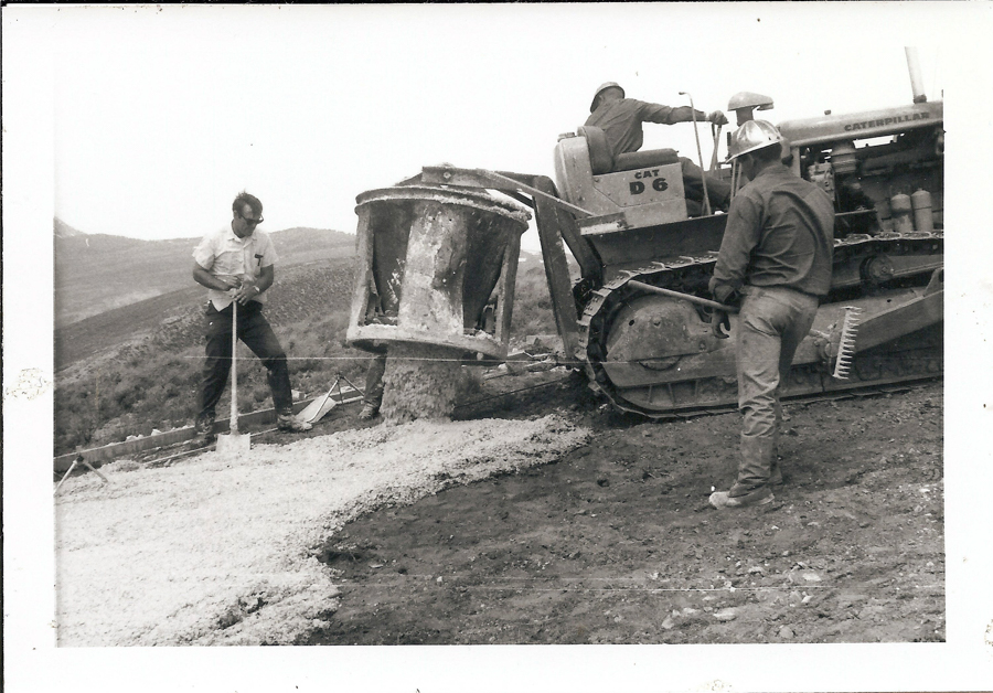 Three men are working on a construction site in a hilly area. One man is operating a Caterpillar bulldozer with "CAT D6" marked on it. Another man is standing and holding a shovel. The third man is wearing a hard hat and appears to be observing or supervising. There is a large container attached to the bulldozer, pouring material onto the ground.