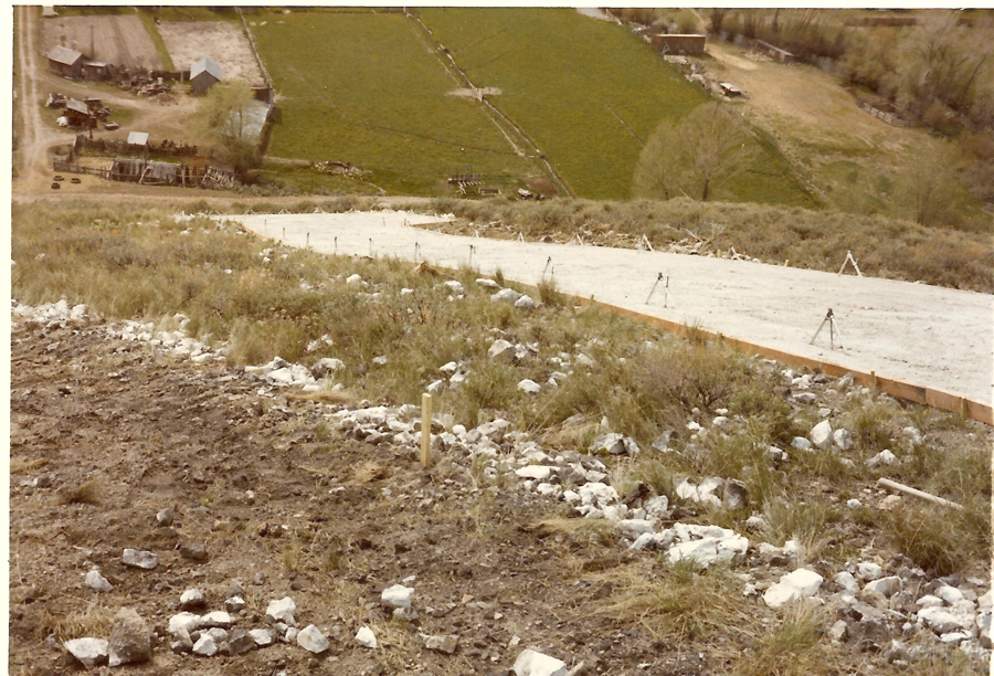 A slope with a mix of grass, rocks, and dirt, leading down to a partially constructed path or road supported by wooden frames and tripods. In the background, there are fields divided by fences, with a few small buildings scattered across the landscape.