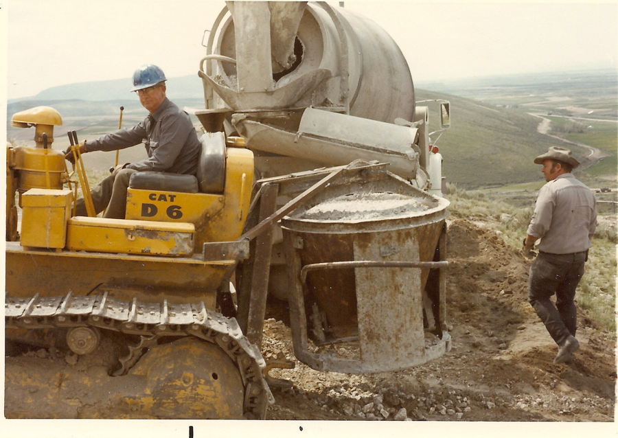A man wearing a blue hard hat is seated on a yellow Caterpillar D6 bulldozer. Another man in a cowboy hat stands nearby, observing. They are working with a large cement mixer on a hilly construction site with a view of a valley and distant landscape in the background.