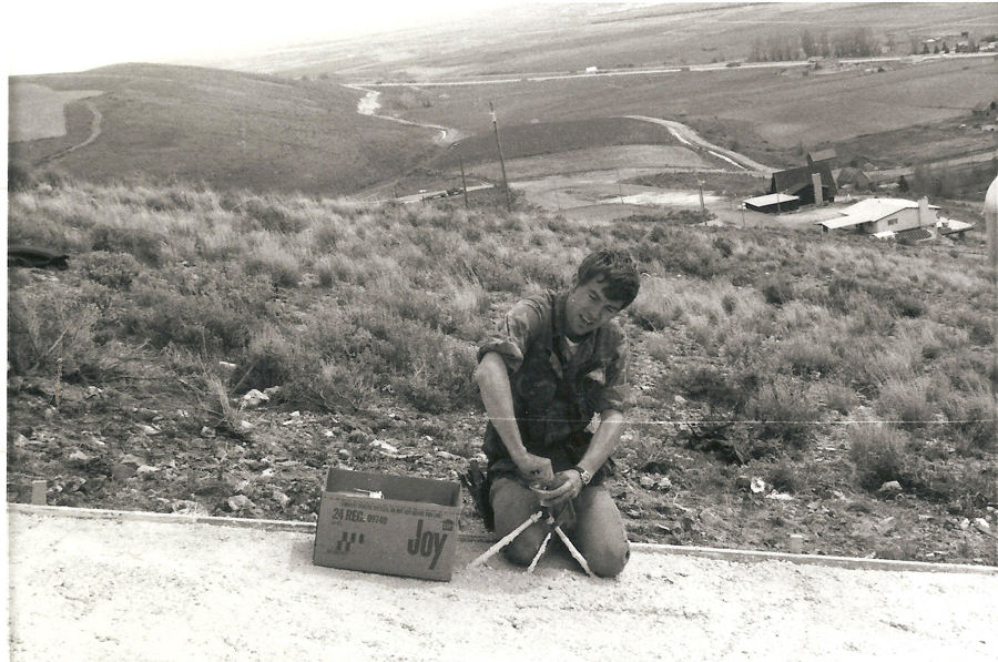 A person is kneeling on the ground, working with a tool on a rocky surface. Next to them is a cardboard box labeled "Joy" with additional text "24 REG. 00140" on it. In the background, there are hills and a few buildings visible in the landscape.