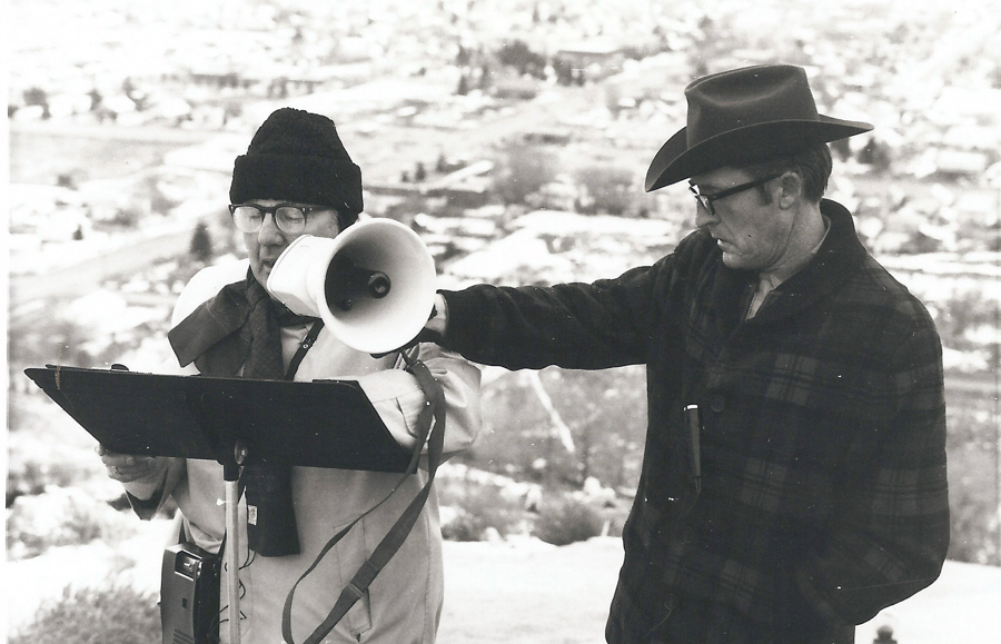 Two individuals are outdoors. One is holding a clipboard and wearing a hat and glasses. The other, also wearing glasses and a hat, is holding a megaphone and appears to be speaking. The background shows a blurred landscape.