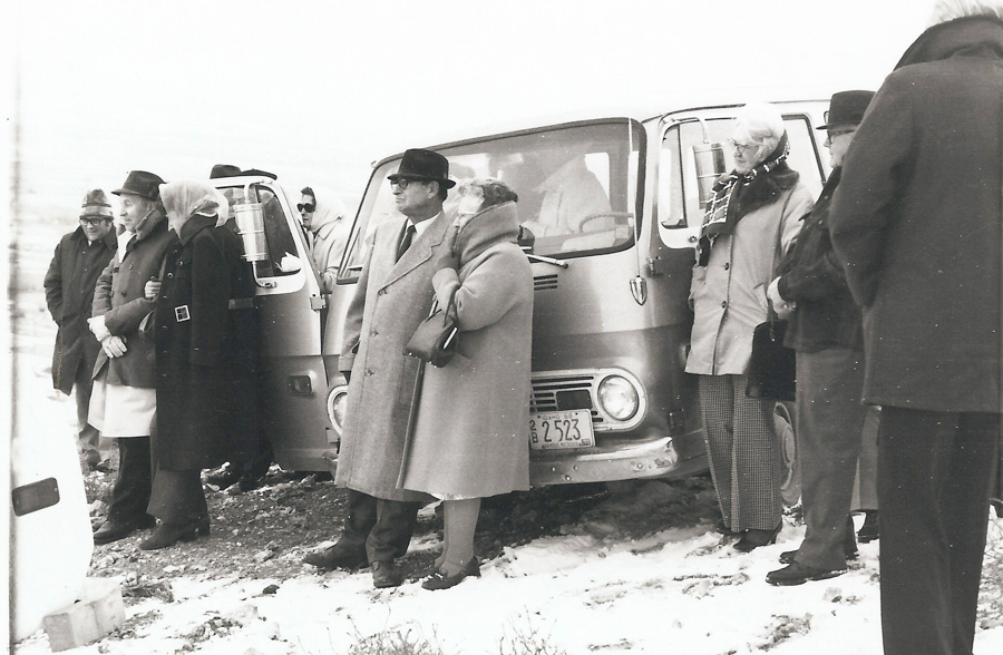 A group of people standing on a snowy ground next to a van. The individuals are wearing winter coats, hats, and scarves. A license plate on the van reads "3B 2523". Some people are standing close to the vans, and one person is seen partially inside the vehicle.