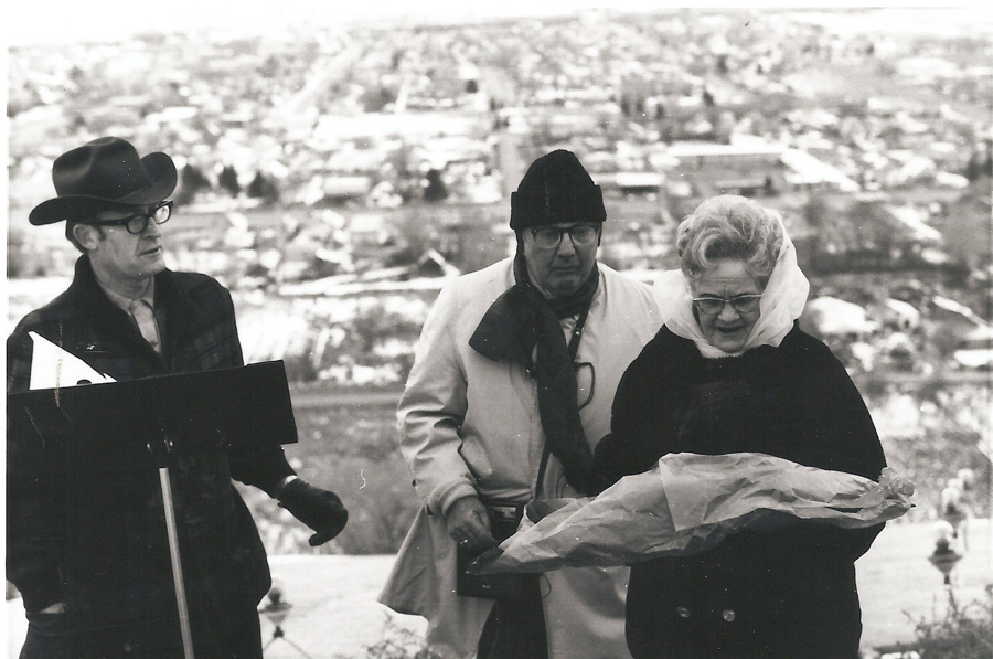 Three people standing outdoors with a view of a city in the background. The person on the left is wearing a hat and glasses, holding a folder or board. The person in the middle is wearing a hat and scarf, with glasses. The person on the right, a woman, is wearing glasses and a headscarf, holding a bouquet wrapped in paper.