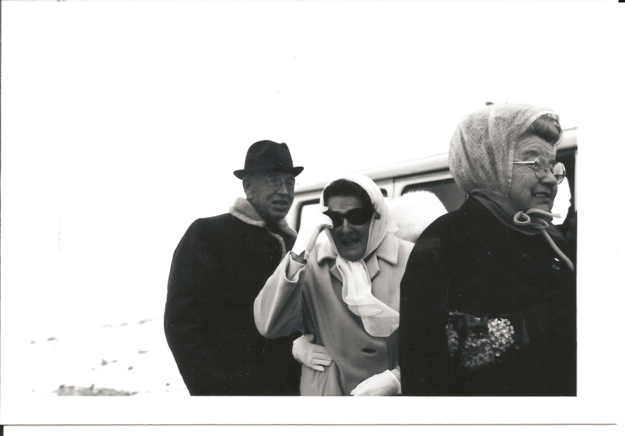 Three people are standing outside. The person on the left is wearing a hat and coat with a fur collar. The person in the middle is wearing sunglasses, a headscarf, a coat, and gloves. They are holding onto the arm of the person on the left. The person on the right is wearing a headscarf, glasses, and a coat with a floral design. A vehicle is partially visible in the background.