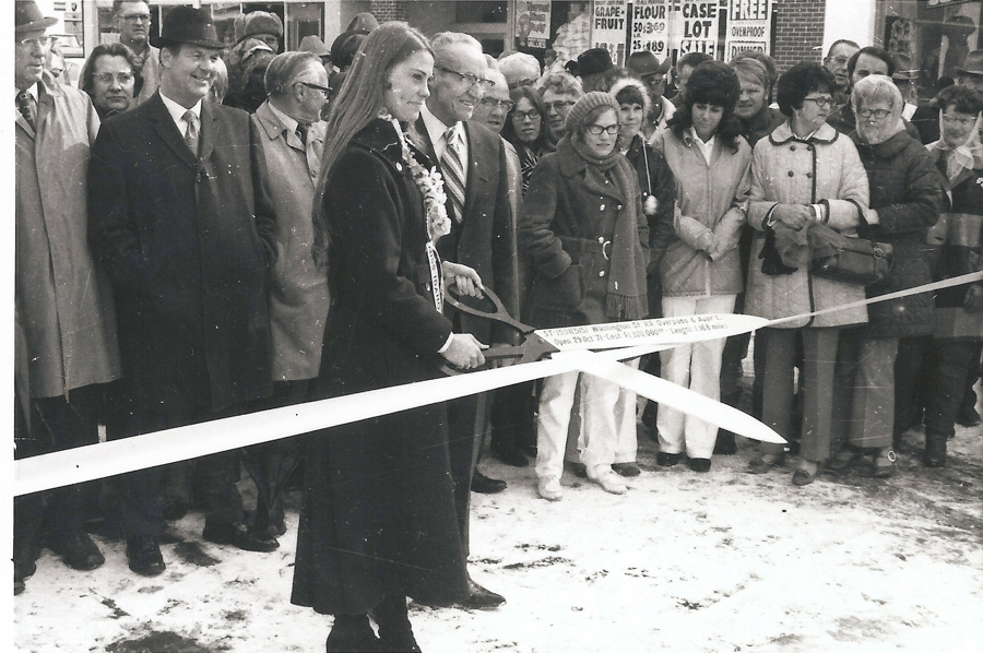 A group of people is gathered outdoors for a ribbon-cutting ceremony. A woman with long hair, adorned with a flower necklace, holds a pair of large scissors to cut the ribbon. Several people stand in the background, wearing coats and hats. There are signs in the background displaying prices and promotions for items like grapefruit and flour.