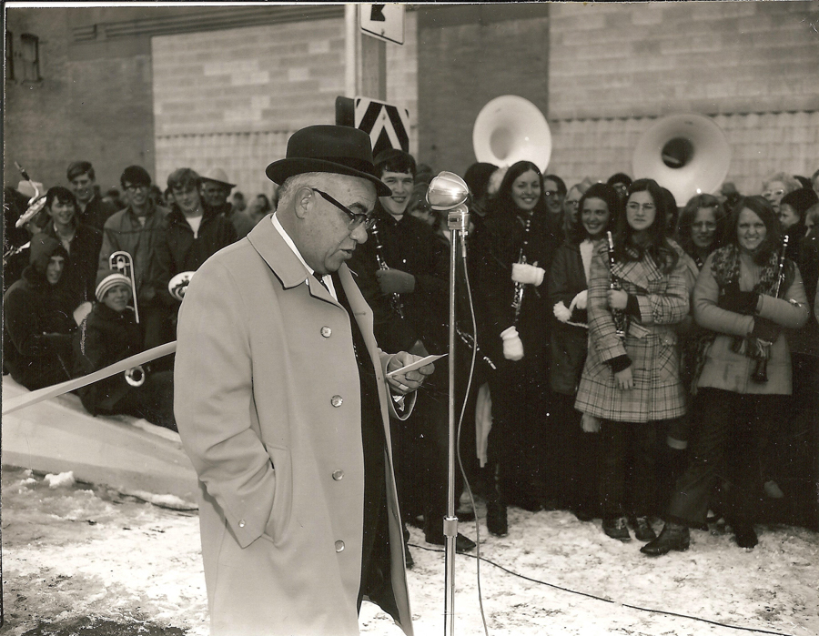 A man wearing a coat and hat stands at a microphone, reading from a piece of paper. Behind him, a group of people, including some holding musical instruments like clarinets and tubas, stand together. The ground is covered in snow, and a building is visible in the background.