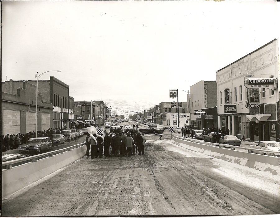 A crowd of people gathered on a street lined with buildings and parked cars. A group with musical instruments, including a tuba, is visible at the forefront. The buildings have various signs, such as "PHONES," "BAR," "CHEVROLET TAPPE CHEVROLET," "BUICK," and "LOANS." Snow is visible on the ground and distant hills. The street features a barrier in the center.