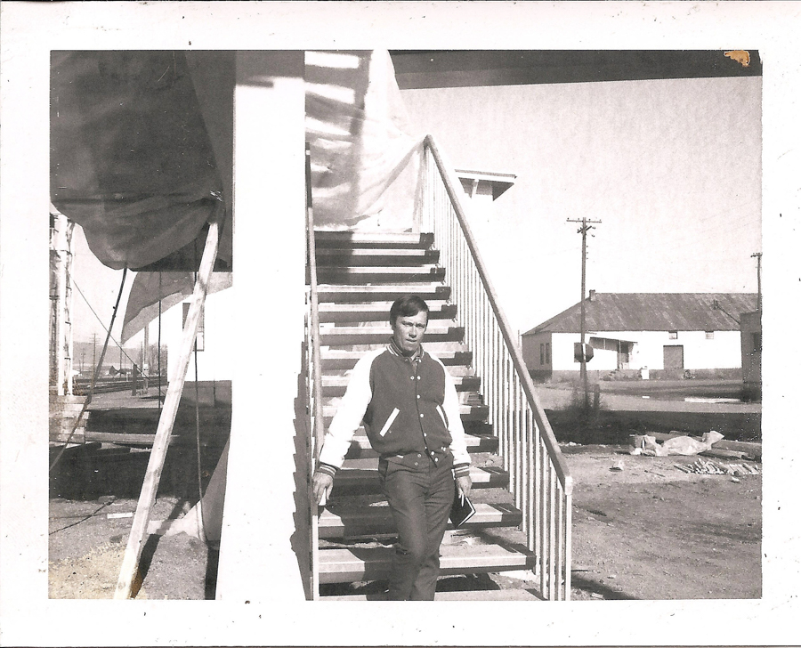 A person standing on an outdoor staircase, wearing a jacket. The staircase is attached to a building, and there are additional buildings in the background, along with telephone poles and an open area with some debris or materials on the ground.
