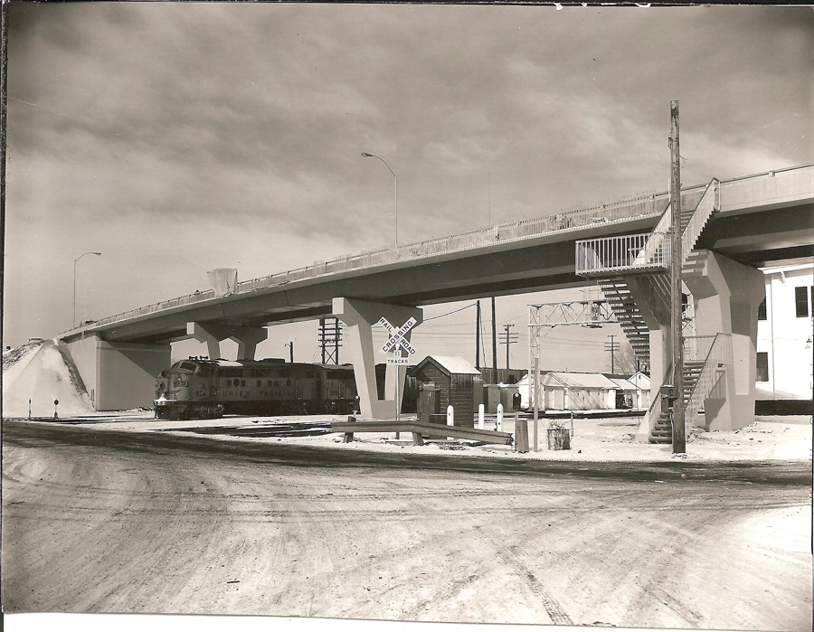 A train labeled "Union Pacific" travels beneath a large overpass. The area is snowy with visible tracks. To the right, there's a staircase leading up to the bridge. A railroad crossing sign reads "Railroad Crossing 5 Tracks." In the background, there are several utility poles and buildings.