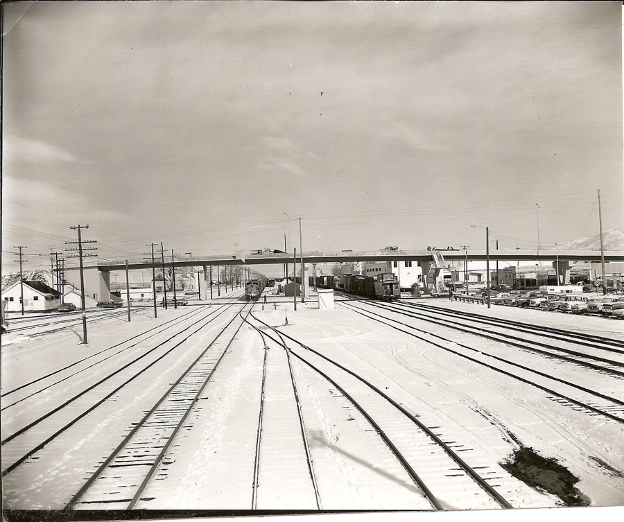 Multiple railroad tracks stretch into the distance in a snowy landscape. A bridge crosses over the tracks, and several train cars are visible beneath it. Buildings and parked cars line the side of the tracks. Utility poles and wires run alongside.