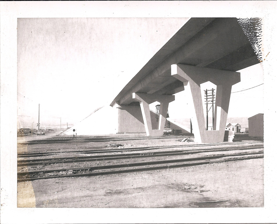 A large bridge structure with triangular supports extends above several sets of train tracks. A small road sign is visible near the tracks. In the background, there are utility poles, a few small buildings, and mountains on the horizon.