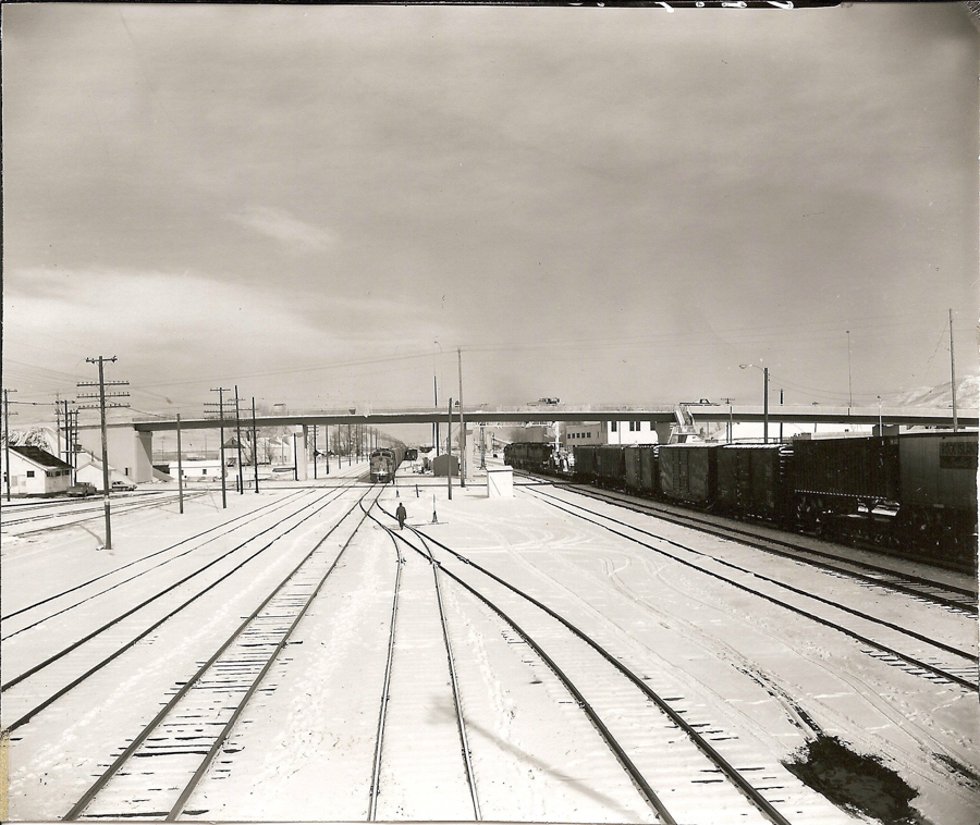 Several railway tracks extending into the distance, with a person walking along one of the tracks. Train cars are visible on some of the tracks. Utility poles line the area, and an overpass is in the background. Snow covers the ground.