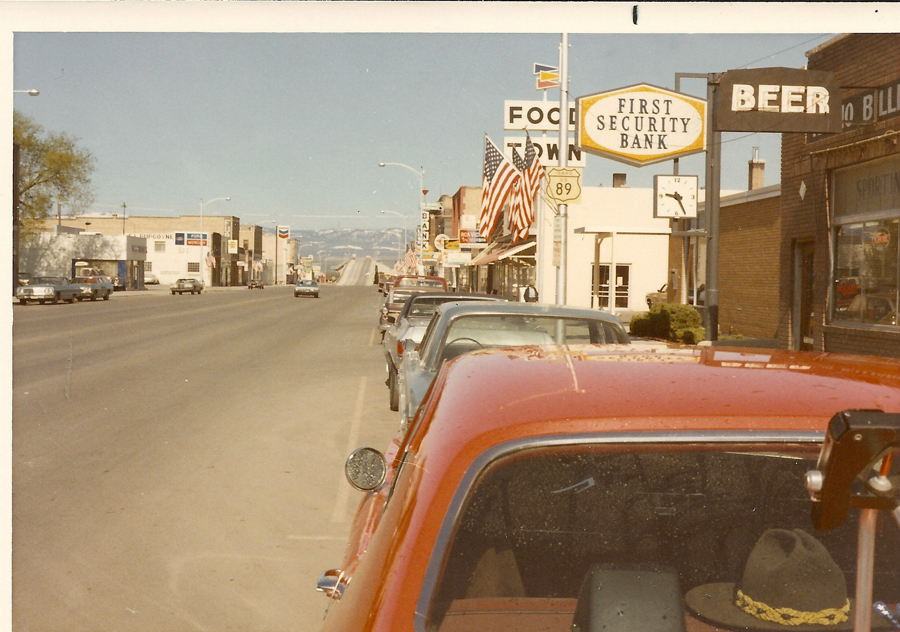A street lined with parked cars on the right side and buildings with various signs. Visible signs include "FOOD TOWN," "FIRST SECURITY BANK," and "BEER." American flags are displayed along the sidewalk. In the distance, cars drive on the road, and a distant view of mountains can be seen. A hat is visible on the back dash of the nearest car.
