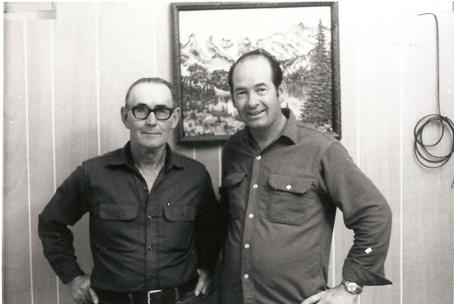 Two men standing side by side in a room. One man is wearing glasses and both are wearing long-sleeved shirts with pockets. Behind them, there is a framed landscape painting of mountains and trees. A coiled wire is hanging on the wall to the right.