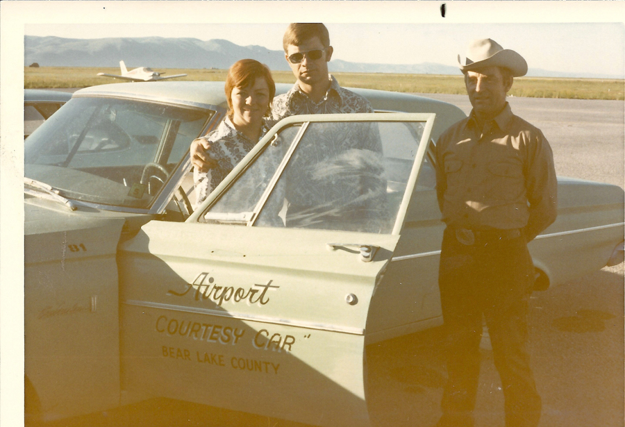 Three people stand next to a car. The car door is open, displaying the text "Airport Courtesy Car Bear Lake County." A man and a woman are standing together by the open door, while another man in a cowboy hat stands to the side. There is an airplane in the background, with mountains visible in the distance.