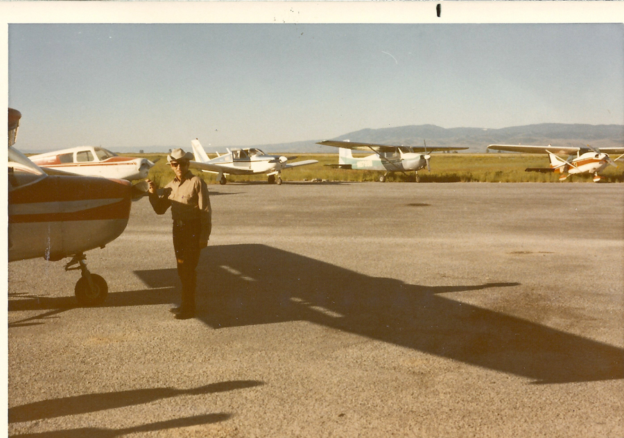 A man wearing a hat, standing on an airfield next to a small aircraft. Several other small airplanes are parked in the background, with mountains visible in the distance.