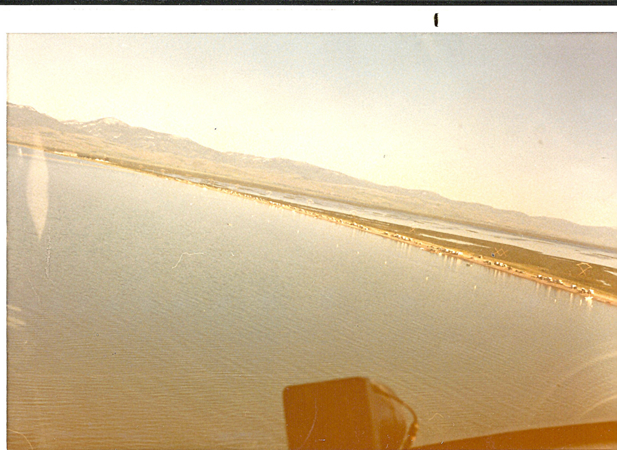Aerial view of a large body of water with a narrow strip of land. Mountains are visible in the distance. Part of an aircraft's interior, possibly the dashboard, is in the foreground. The view appears to be slightly tilted.