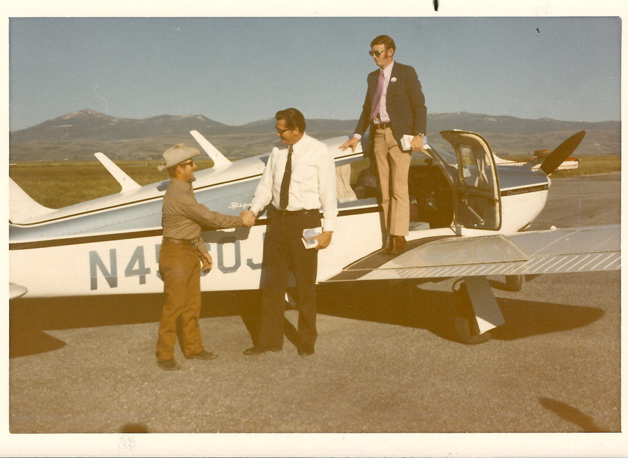Two men are standing next to a small aircraft, shaking hands. One man is wearing a hat and casual clothing, while the other is in a shirt and tie. A third man, wearing a suit and holding a book, stands on the wing of the plane. The plane has "N4509J" written on its side. A mountainous landscape is visible in the background.