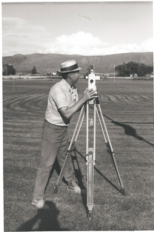 A man is standing on a grassy field using a theodolite mounted on a tripod. He is wearing a short-sleeved shirt, pants, and a straw hat. The background includes hills and trees with a few structures visible in the distance.