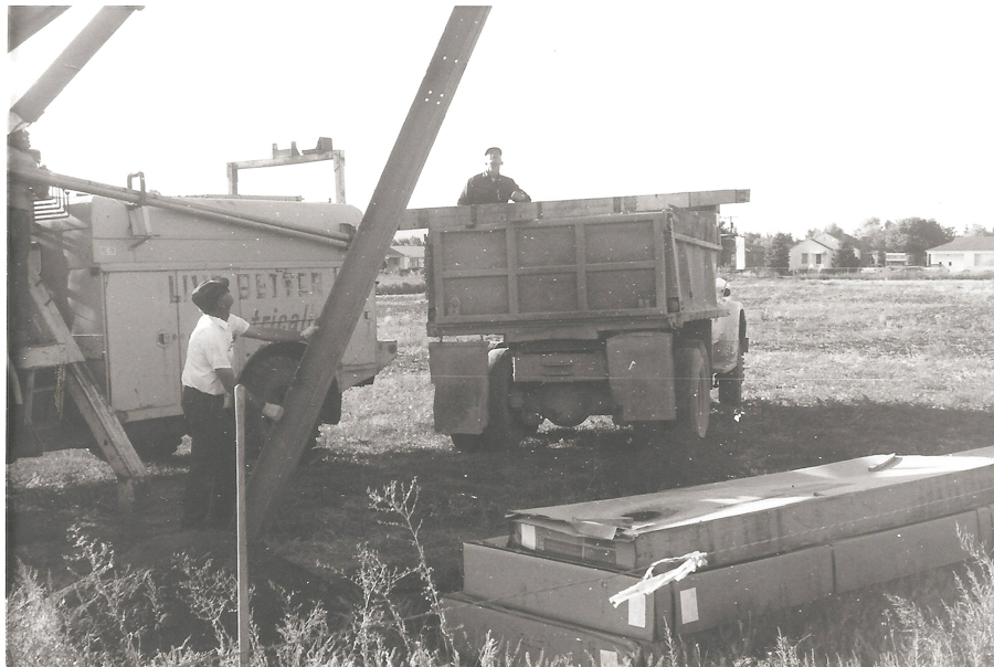 Two men are working in a field. One man stands beside a truck with a large chute, and another is in the back of a separate truck. The trucks are positioned on a grassy area with some buildings visible in the background. There is a stack of boxes on the ground nearby. The truck with the chute has partial text visible on it.